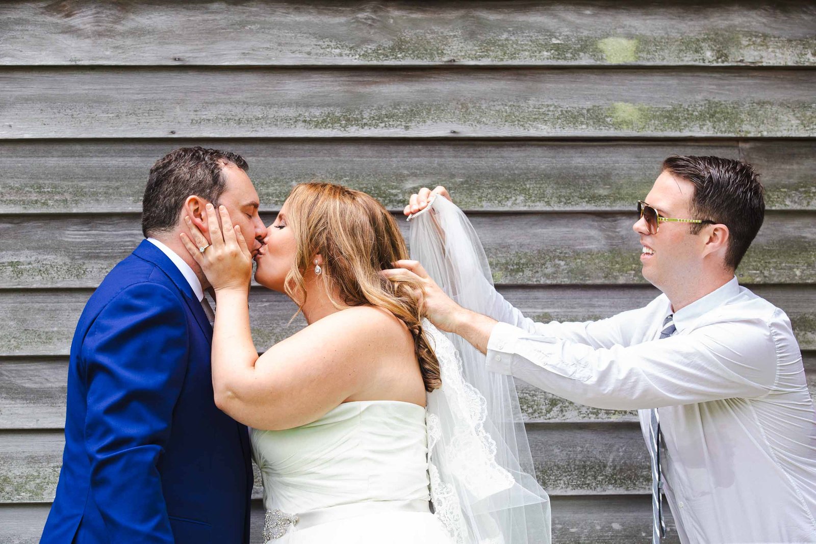 bride and groom kiss while friend adjusts veil