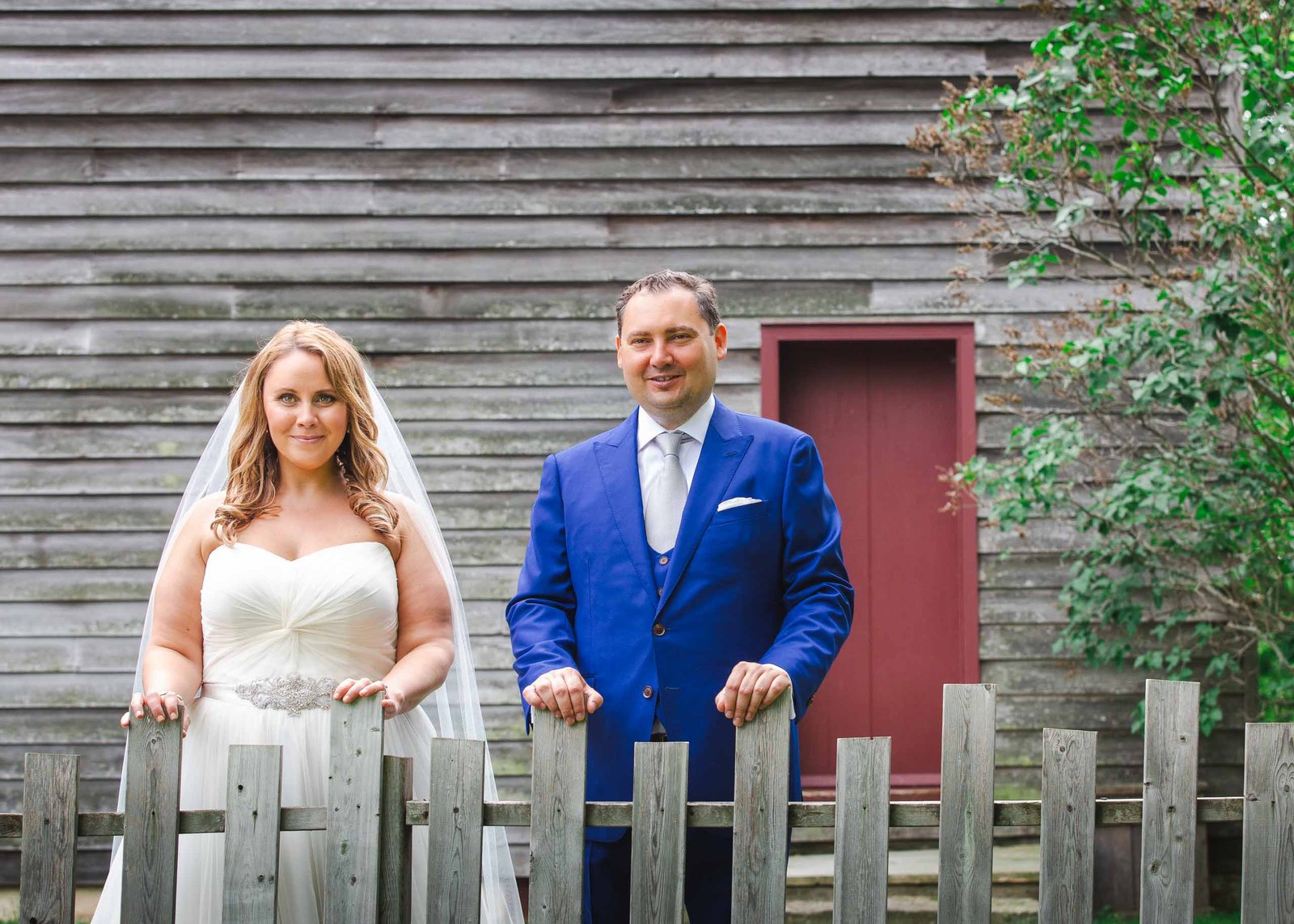 bride and groom stand at a fence in front of a barn